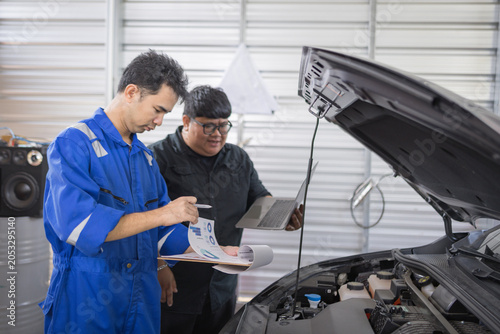 Two mechanics examine a car engine together. One holds a clipboard, the other a laptop. They focus on the engine's components closely. Their expressions show concentration and teamwork