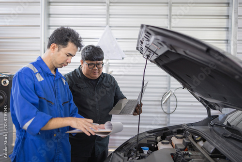Two mechanics examine a car engine together. One holds a clipboard, the other a laptop. They focus on the engine's components closely. Their expressions show concentration and teamwork