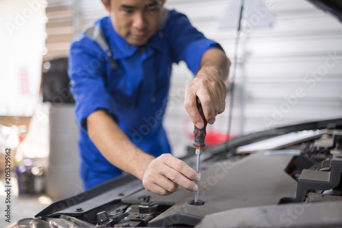 Two mechanics examine a car engine together. One holds a clipboard, the other a laptop. They focus on the engine's components closely. Their expressions show concentration and teamwork