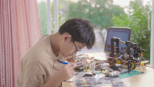 Asian young student using a soldering iron for electronics and robotics projects at home with robotic equipment and a laptop. Concept of STEM education, engineering skills, and innovation.