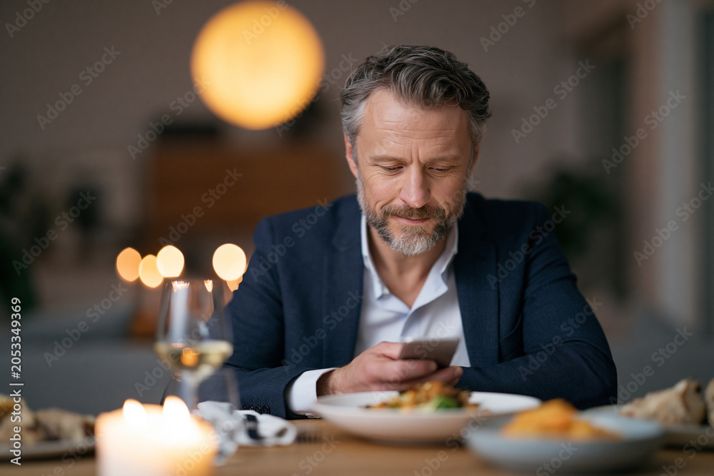 Fototapeta premium Man sits at dinner, smiling, scrolling phone. Candlelight glows softly on wooden table. Wine glass, plates, and appetizers surround him. Warm lighting and cozy background create intimate mood