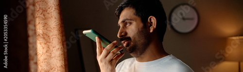 Foto Header of young adult Indian man using smartphone at home, speaking into screen and engaging in prolonged device use, suggesting digital dependence