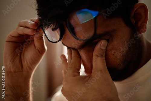 Close up of young adult man rubbing eyes and removing glasses at home, showing digital fatigue and screen overuse. Useful for wellness campaigns, tech burnout content