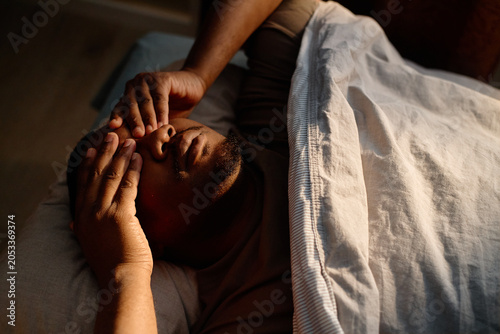 Obraz na płótnie High angle view of young adult Black man lying in bed, covering eyes and struggling with fatigue and sleep disruption at home