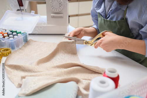 Woman's hands cutting fabric with golden scissors on white table with sewing machine and thread spools, a sewing machine, a box of colorful thread spools