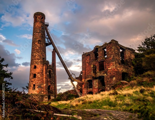 Ruins of an old industrial structure with a tall brick tower and dilapidated building under a dramatic, cloudy sunset
