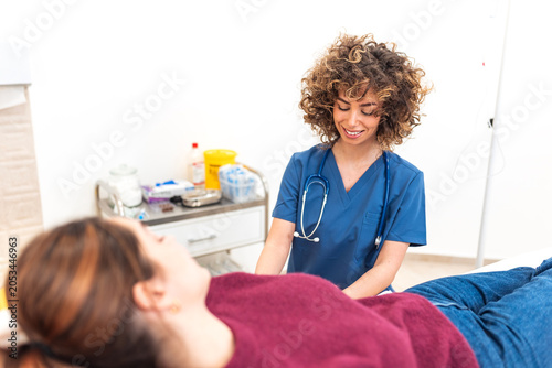 Smiling nurse in blue scrubs preparing female patient for intravenous infusion therapy in clinic room. Professional medical worker checking woman lying on examination bed for iv drip treatment.