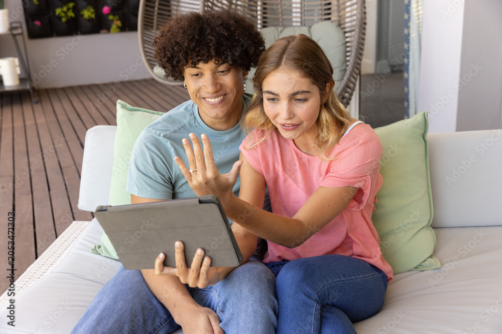 Fototapeta premium Couple sitting on cushioned sofa on deck holding gray tablet gesturing in t shirts and jeans