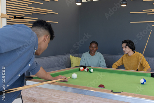 Three male friends playing pool on green felt table in lounge corner, watching balls, holding cues