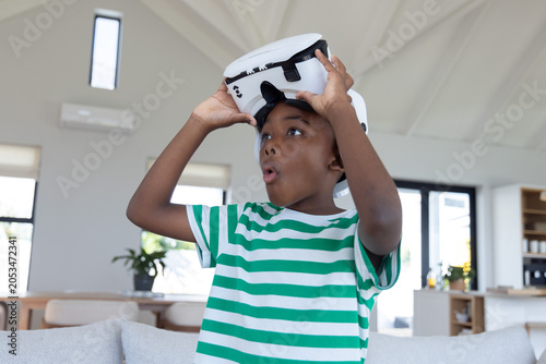 Canvas Print African American child lifting white VR headset behind gray couch at home in gre
