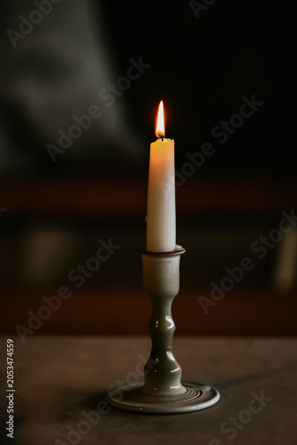 A single white lit candle in a ceramic holder casting a warm glow against a dark, rustic background.