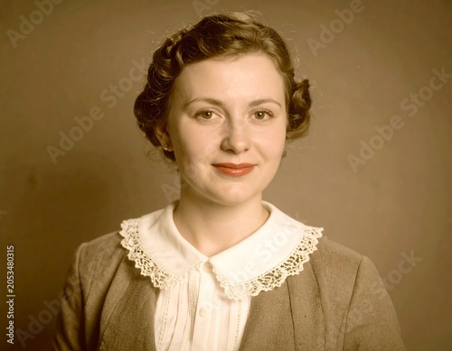 Portrait of a woman with curly hair, wearing a white collared blouse, grey jacket, and red lipstick. Her expression is calm