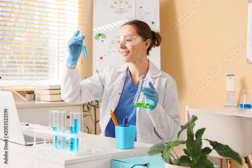 Female scientist in lab coat and safety glasses examining test tube with blue...