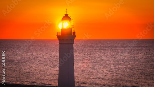 AERIAL  LENS FLARE  CLOSE UP  Lighthouse tower with the setting sun shining behind the lantern room over the Mediterranean Sea. Warm golden light colours the sky  while horizon stays crisp and calm.