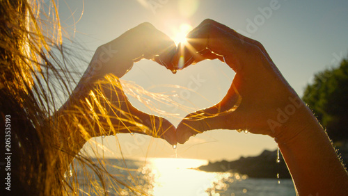 Photography CLOSE UP, LENS FLARE, SILHOUETTE: Young woman makes a heart gesture toward the setting sun as water drips from her hands