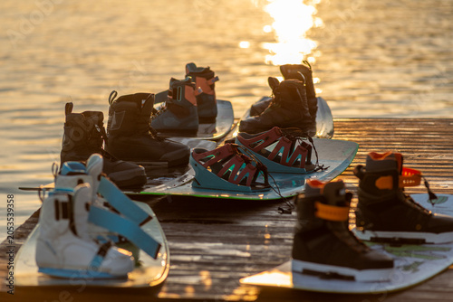 Wakeboard boots and boards lying on a wooden dock near calm water at sunset, suitable for summer sport, outdoor recreation, lake vacation, active lifestyle, and water adventure concepts