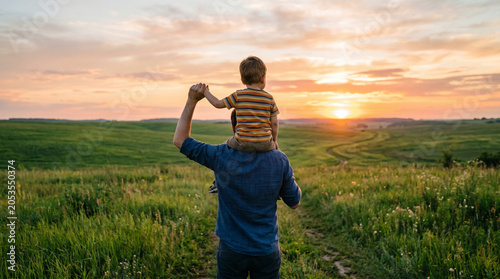 Back view of loving father carrying his little son on shoulders while watching beautiful sunset over green field, holding hands in warm evening light and creating happy childhood family memories