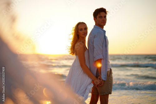 Romantic couple holding hands on the beach at sunset with ocean waves and golden glow. Generative AI