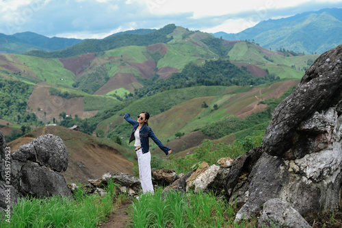 Bild auf Leinwand A woman stands joyfully on a rocky hill overlooking lush, patchy green mountains under a cloudy sky