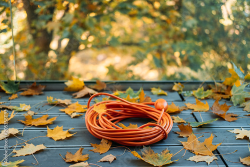 orange coiled extension cord lies on gray ribbed wooden decking next to scattered yellow autumn maple leaves. Outdoor cable insulation, waterproof electrical equipment, extension cord safety.