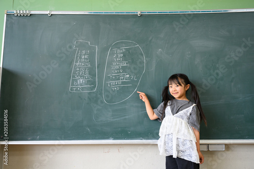 教室の黒板に書かれた時間割を指差して笑顔を見せる女子小学生