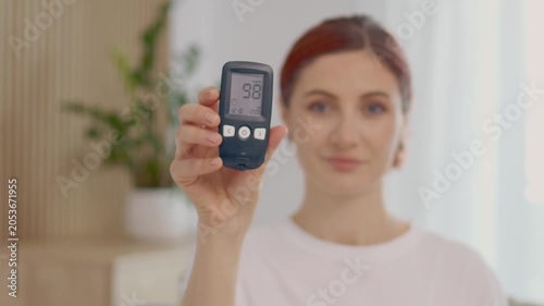 A woman holds a glucometer up to the camera, showing a reading of 98 mg dL. She is in a home environment with plants in the background.