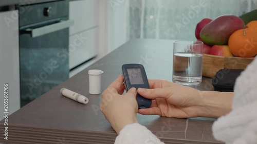 A woman checks her blood sugar results using a glucometer diary while sitting at the kitchen table. She reviews the history of her glucose levels carefully.