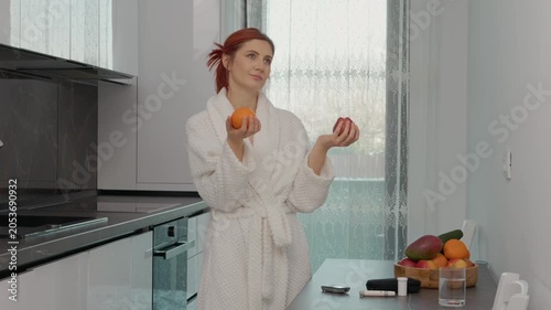 A woman stands in her kitchen holding an apple and an orange while looking at her glucometer. She is deciding which fruit to eat after checking her blood sugar level.
