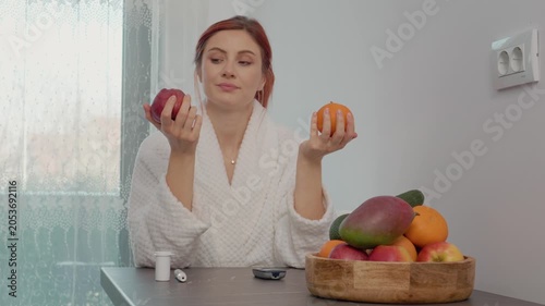 A woman stands in her kitchen holding an apple and an orange after checking her blood sugar level. She considers which fruit to eat for her health.