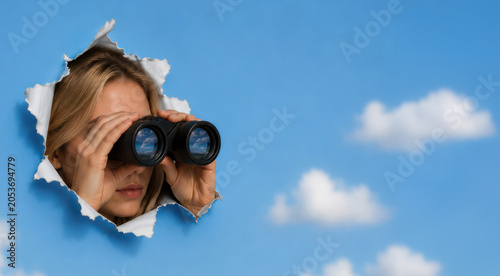 Creative conceptual image of a woman peeking through torn blue paper while looking through binoculars against a sky-themed background with clouds. Modern business and lifestyle concept symbolizing exp
