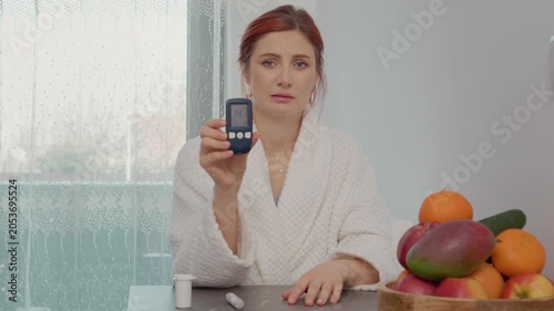 A woman sits in her kitchen wearing a robe. She shows a glucometer reading of 141 mg dL, indicating a high blood sugar level. She looks concerned about her health.