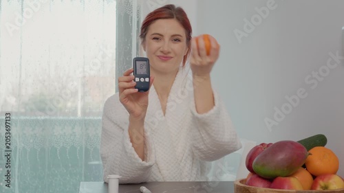 A woman measures her blood sugar level with a glucometer in the kitchen. The reading shows 87 mg dL. She is happy to eat an orange as part of her snack.