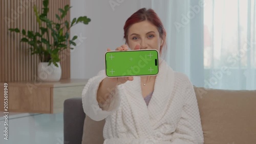 A woman displays a smartphone with a green screen in her home. The setting has natural light and a plant, creating a simple environment.