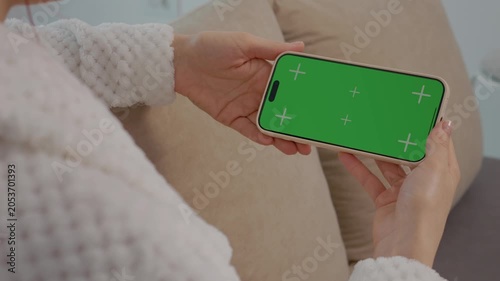 A woman sits on a couch at home and holds a smartphone with a green screen. She looks at the phone while relaxing in her comfortable space.