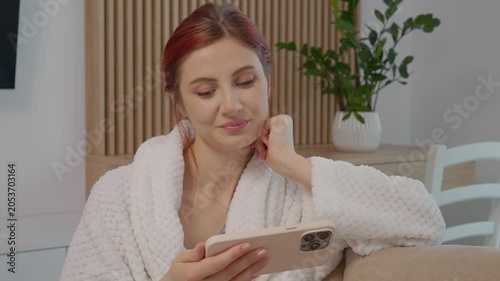 A woman is on a sofa in her living room. She holds a smartphone in her hand and focuses on the screen. The atmosphere is casual and relaxed.