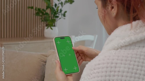 A woman sits on a sofa in the living room. She looks at a phone with a green screen. The space is cozy and has plants in the background.