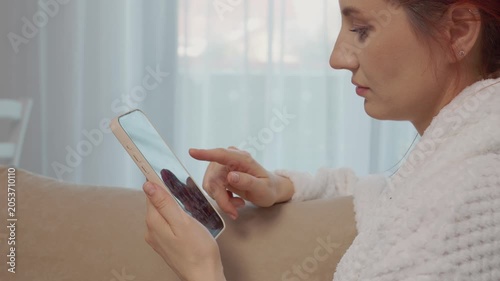 A woman sits on a couch at home, holding a smartphone. She swipes on the screen while engaging with social media and connecting with friends.