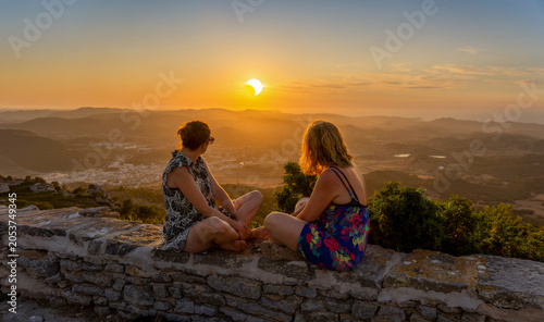 Konstfotografi Two young female friends in summer dresses sitting on a stone wall watching a spectacular sunset during a solar eclipse from the Monte Toro viewpoint in Menorca, Spain