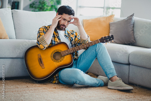 Tableau sur toile Young man with acoustic guitar sitting on living room floor, rubbing his temples