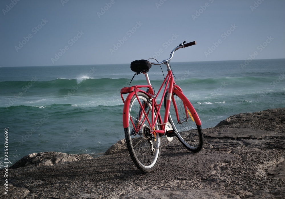 Fototapeta premium Bright red vintage bicycle parked on a rocky stone ledge overlooking the calm blue ocean waves