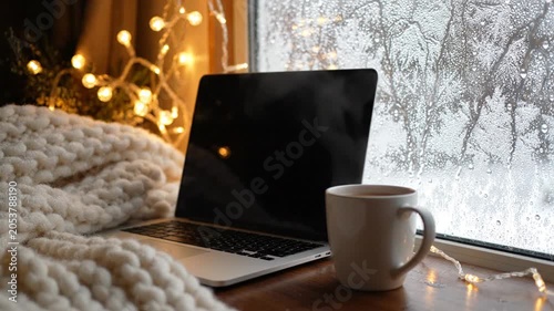 Cozy winter scene with laptop, warm drink, knitted blanket, and sparkling fairy lights by a frosted window
