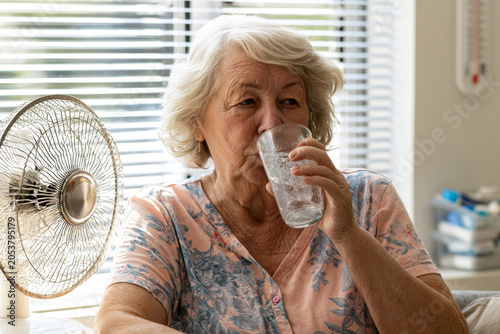 Senior woman drinking water at home near a fan during hot summer weather, illustrating hydration, heatwave safety, and elderly health care