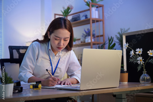 Bild auf Leinwand Asian woman writing in notebook with laptop in office at night for business plan