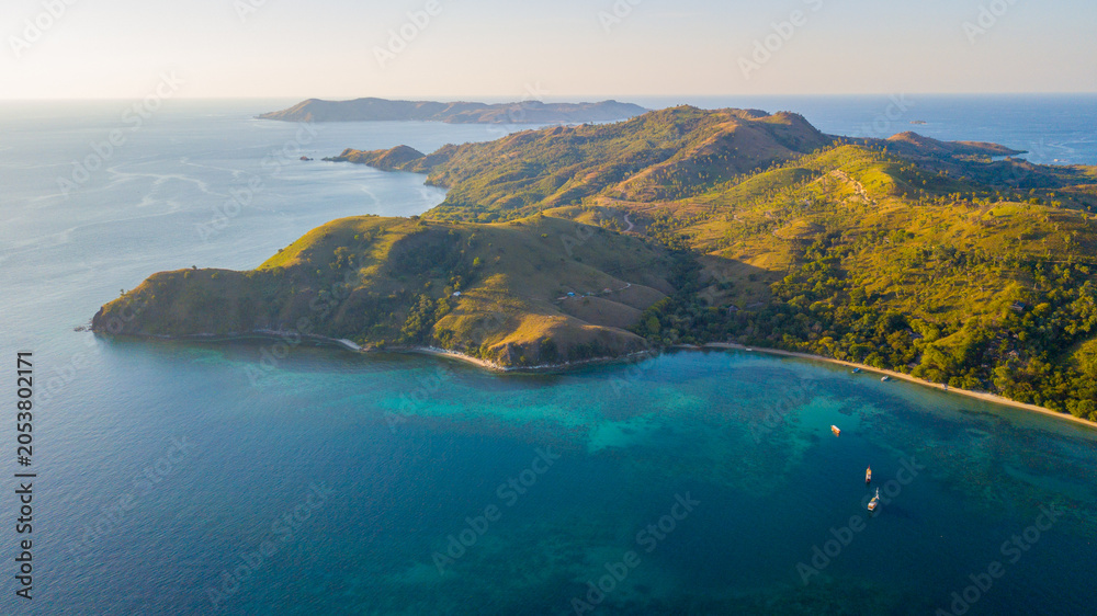 Fototapeta premium Aerial sunset view of tropical rolling hills, turquoise coral reef, and boats in Komodo National Park, Indonesia.