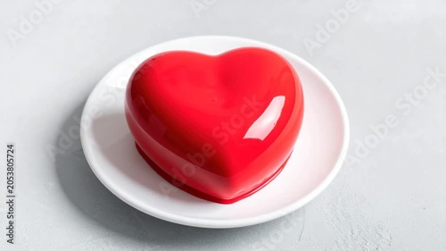 Red heart-shaped dessert with shiny glaze being poured on a white plate against a grey background
