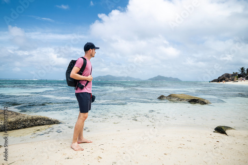 Quadro em tela Male tourist with backpack and cap standing on tropical beach in Seychelles, looking around at scenic landscape