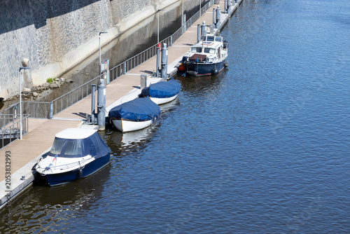 Foto Four boats docked along a wooden pier beside a calm river, with a stone wall and
