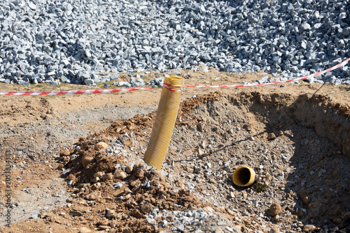 Yellow drainage pipe protruding from a construction site trench surrounded by gravel and soil, with safety tape marking the area for caution