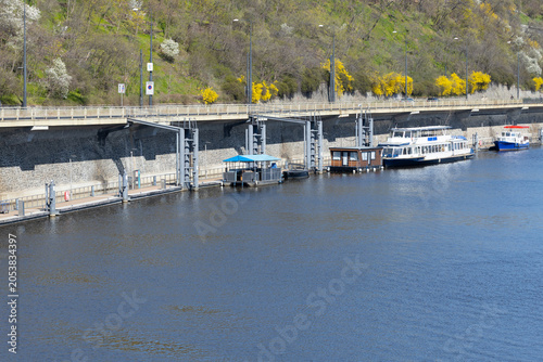 Scenic riverside dock with boats moored along the waterfront, surrounded by lush greenery and blooming trees under a clear blue sky in a tranquil urban setting