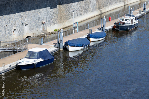Several boats docked along a stone wall at a marina, with calm water reflecting the blue sky and sunlight creating a serene atmosphere